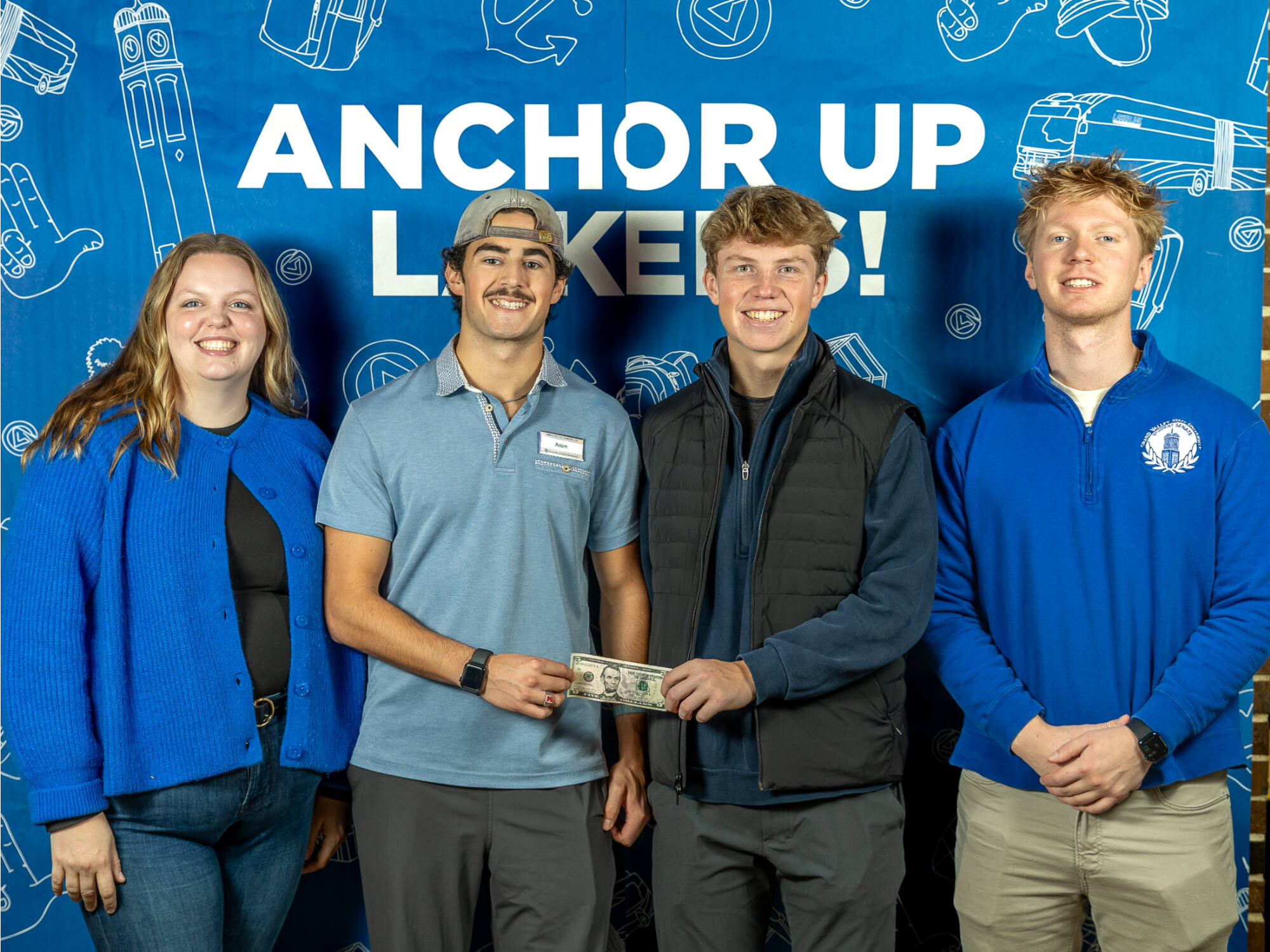 four individuals, one on the left smiling wearing a blue cardigan, two in the middle both holding the same dollar bill, one on the right wearing a gvsu student senate sweater